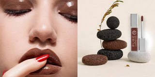 Close-up of a model wearing glossy brown lips with a finger touching the lip, paired with a Pure Cosmetics lip gloss displayed on stacked stones against a neutral background.