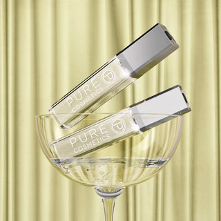 Two Pure Cosmetics Clear Light Up Lip Gloss tubes in a champagne coupe glass against a soft butter-yellow backdrop, showcasing the August trending shade.